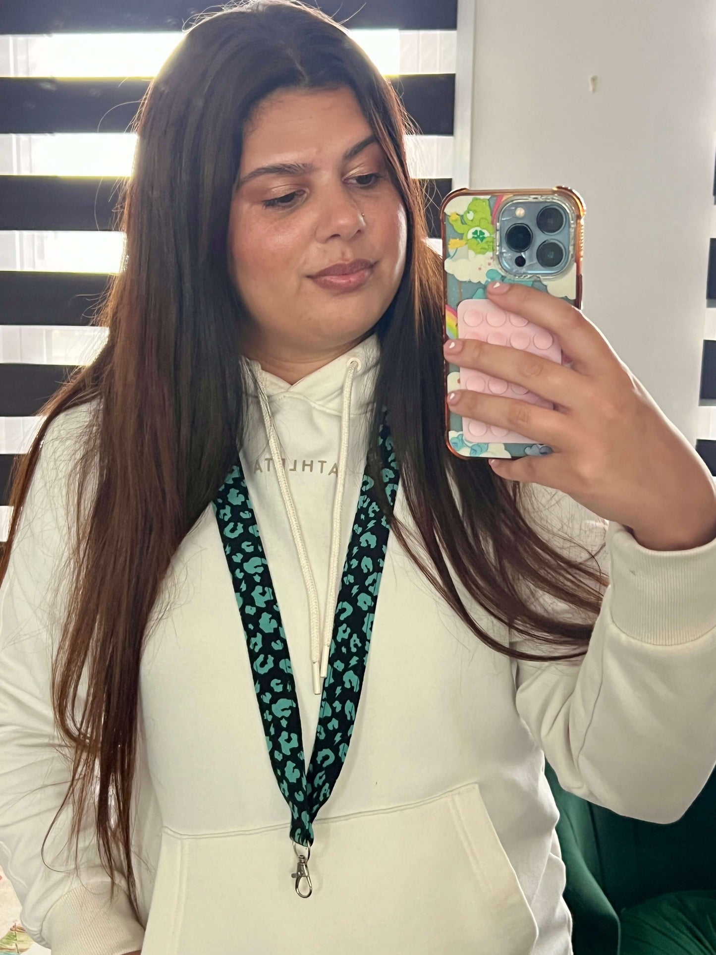 Brand Owner Elana taking a mirror selfie with a phone, wearing a white hoodie and green leopard print lanyard.