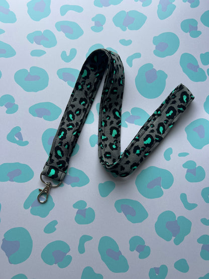 Sew Much Love grey fabric lanyard with a bold snow leopard print in teal and black, featuring a metal D-ring and swivel clasp.